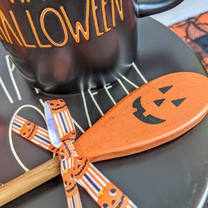 Op de afbeelding: Een zwarte keramische mok met de tekst "Happy Halloween" in oranje letters. De mok staat op een zwart bord met een houten lepel met een gesneden pompoengezicht. De lepel is vastgebonden met een lint met een pompoenpatroon.