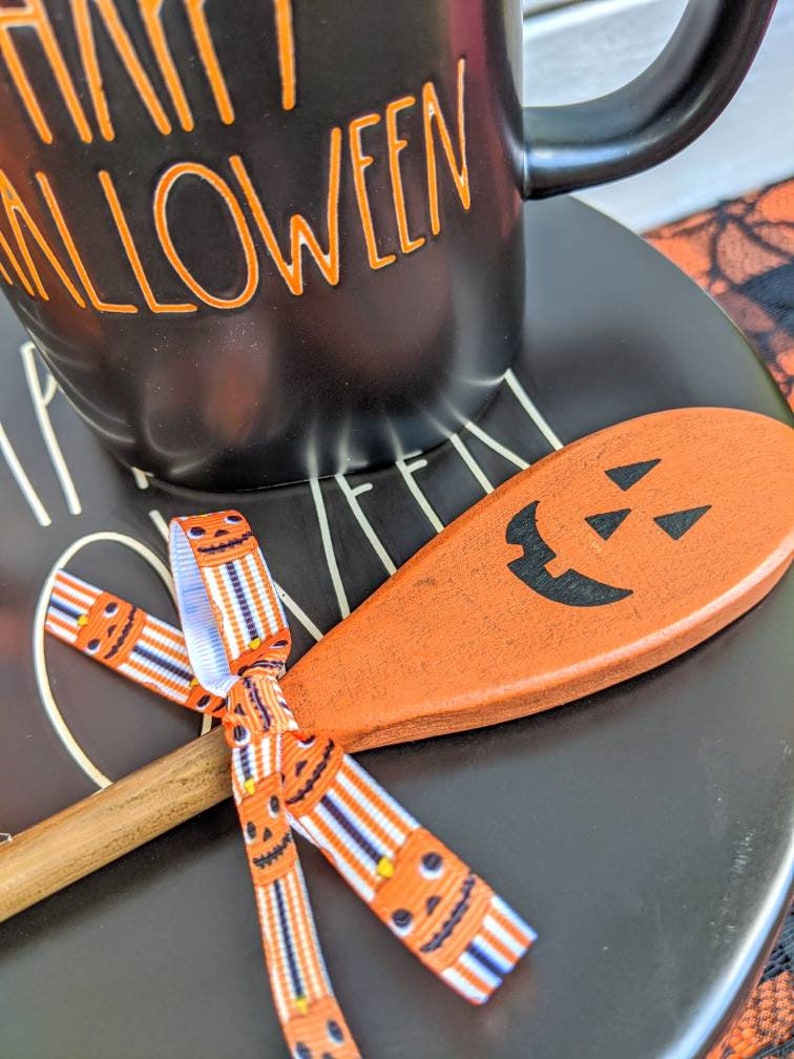 Op de afbeelding: Een zwarte keramische mok met de tekst "Happy Halloween" in oranje letters. De mok staat op een zwart bord met een houten lepel met een gesneden pompoengezicht. De lepel is vastgebonden met een lint met een pompoenpatroon.