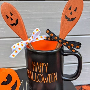 Op de afbeelding: Zwarte keramische mok met oranje "Happy Halloween" tekst, twee oranje houten lepels met zwarte pompoengezichten en oranje-witte snoepmaislint.