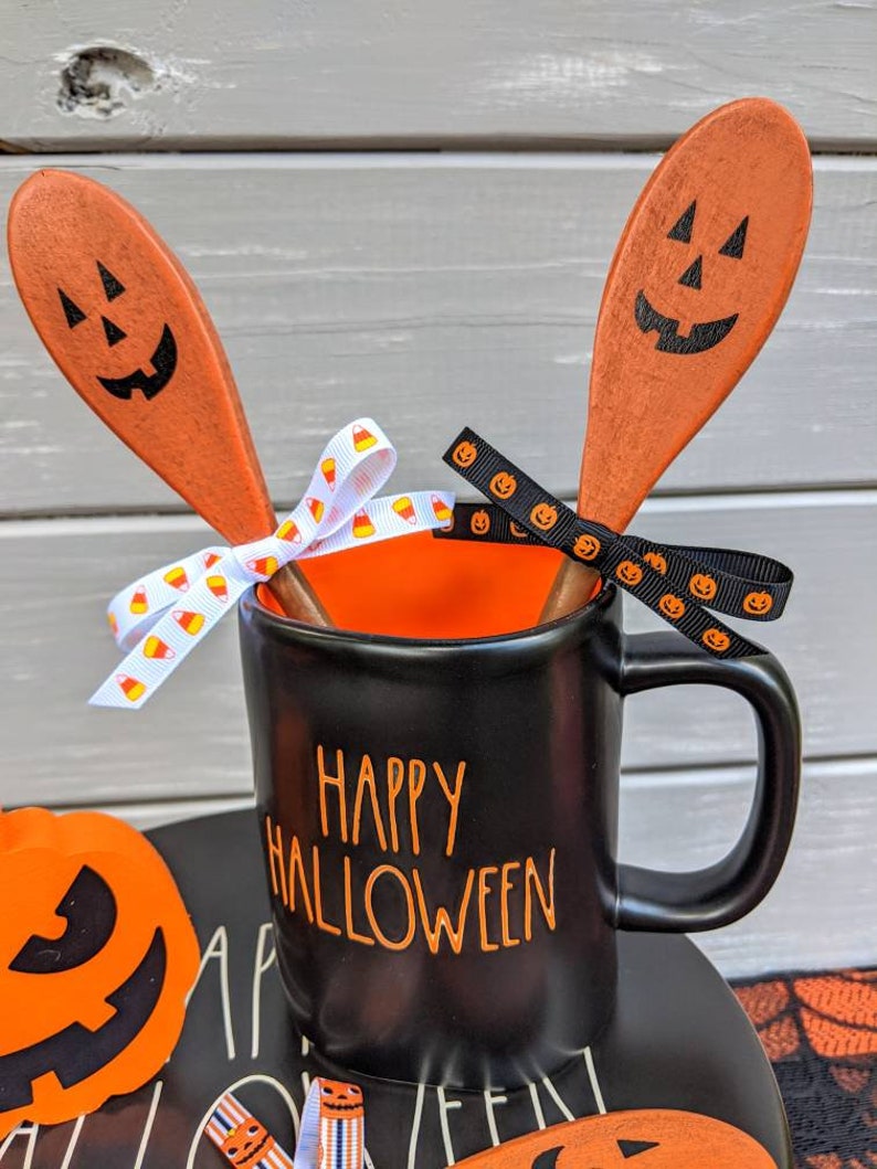 Op de afbeelding: Zwarte keramische mok met oranje "Happy Halloween" tekst, twee oranje houten lepels met zwarte pompoengezichten en oranje-witte snoepmaislint.