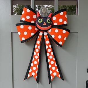 May include: A decorative bow featuring an orange fabric with white polka dots and black glittery edges. Centered on the bow is a black cat face with green eyes, a pink nose, and a wide smiling mouth. The bow has two long hanging tails.