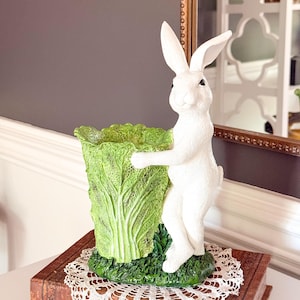 Könnte beinhalten: Eine weiße Hasenfigur, die einen grünen, kohlförmigen Behälter hält. Der Hase steht aufrecht mit aufgestellten Ohren. Der Behälter steht auf einem braunen Buch mit einer weißen Spitzendecke. Der Hintergrund ist eine neutrale graue Wand.