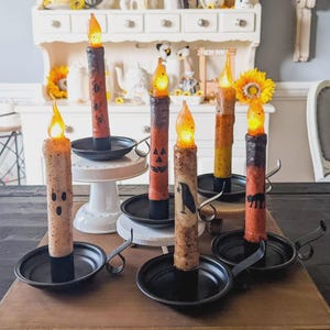 May include: A collection of six battery-operated taper candles with flickering orange flames, each featuring unique Halloween-themed graphics like ghosts, spiders, jack-o'-lanterns, crows, and black cats, set in individual black metal candle holders.
