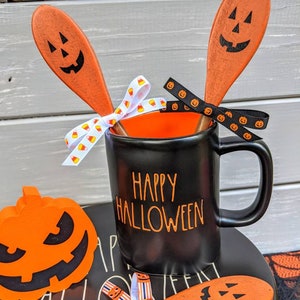 Op de afbeelding: Een zwarte koffiemok met de tekst "Happy Halloween" in oranje letters. De mok is versierd met twee oranje houten lepels die beschilderd zijn met pompoengezichten en vastgebonden met een zwart-wit lint.