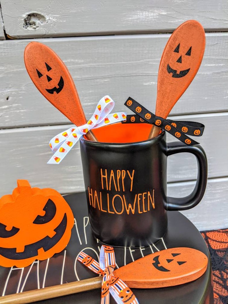 Op de afbeelding: Een zwarte koffiemok met de tekst "Happy Halloween" in oranje letters. De mok is versierd met twee oranje houten lepels die beschilderd zijn met pompoengezichten en vastgebonden met een zwart-wit lint.