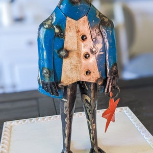 May include: A metal sculpture of a bird-like figure wearing a blue jacket and a white vest. The sculpture has welded details and holds a red star. The figure stands on thin metal legs.