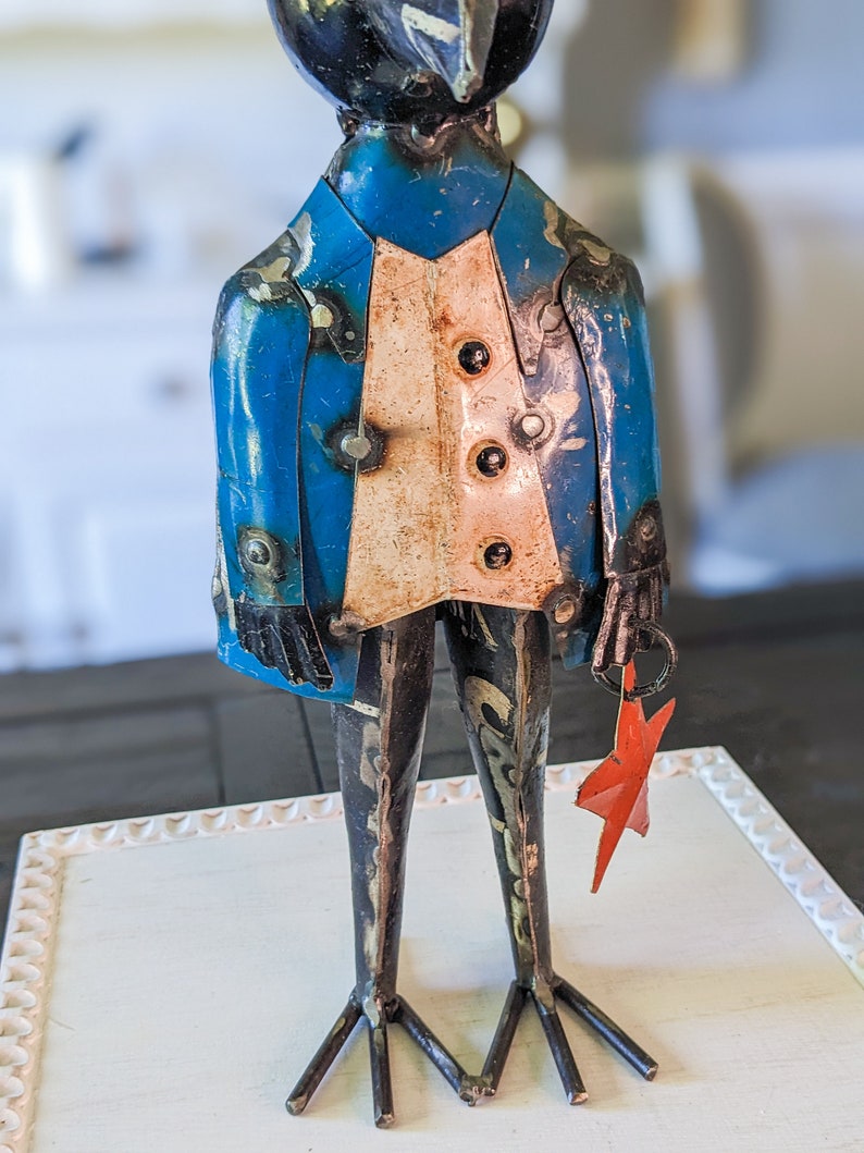 May include: A metal sculpture of a bird-like figure wearing a blue jacket and a white vest. The sculpture has welded details and holds a red star. The figure stands on thin metal legs.