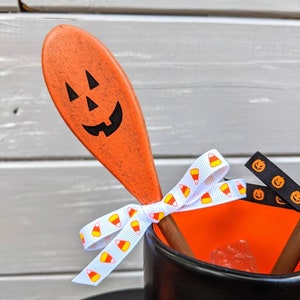 Op de afbeelding: Een zwarte mok met de tekst "Happy Halloween" aan de voorkant. De mok is versierd met een oranje houten lepel die is beschilderd met een pompoengezicht. De lepel is vastgebonden aan de mok met wit lint met een snoepmaispatroon en zwart lint met een pompoengezicht.