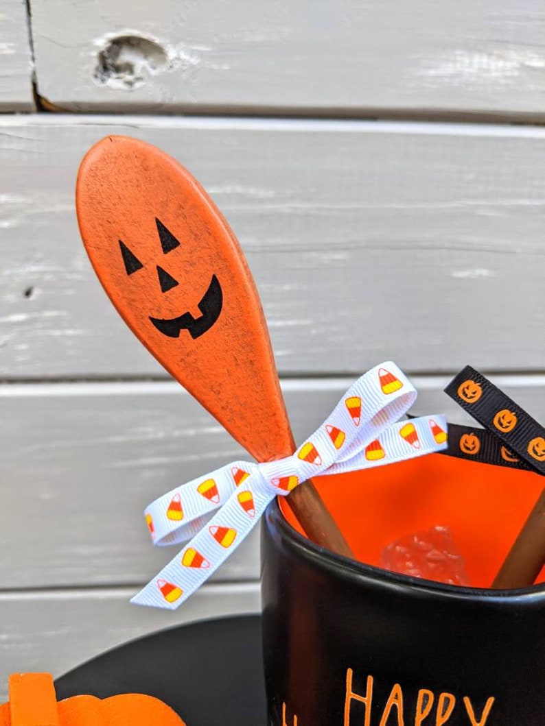 Op de afbeelding: Een zwarte mok met de tekst "Happy Halloween" aan de voorkant. De mok is versierd met een oranje houten lepel die is beschilderd met een pompoengezicht. De lepel is vastgebonden aan de mok met wit lint met een snoepmaispatroon en zwart lint met een pompoengezicht.
