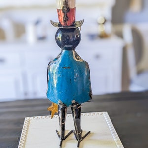 May include: A metal sculpture of a figure with a top hat, blue coat, and star-shaped accessory. The hat is white, red, and silver. The figure is standing on a white, framed surface. The sculpture is a decorative item.