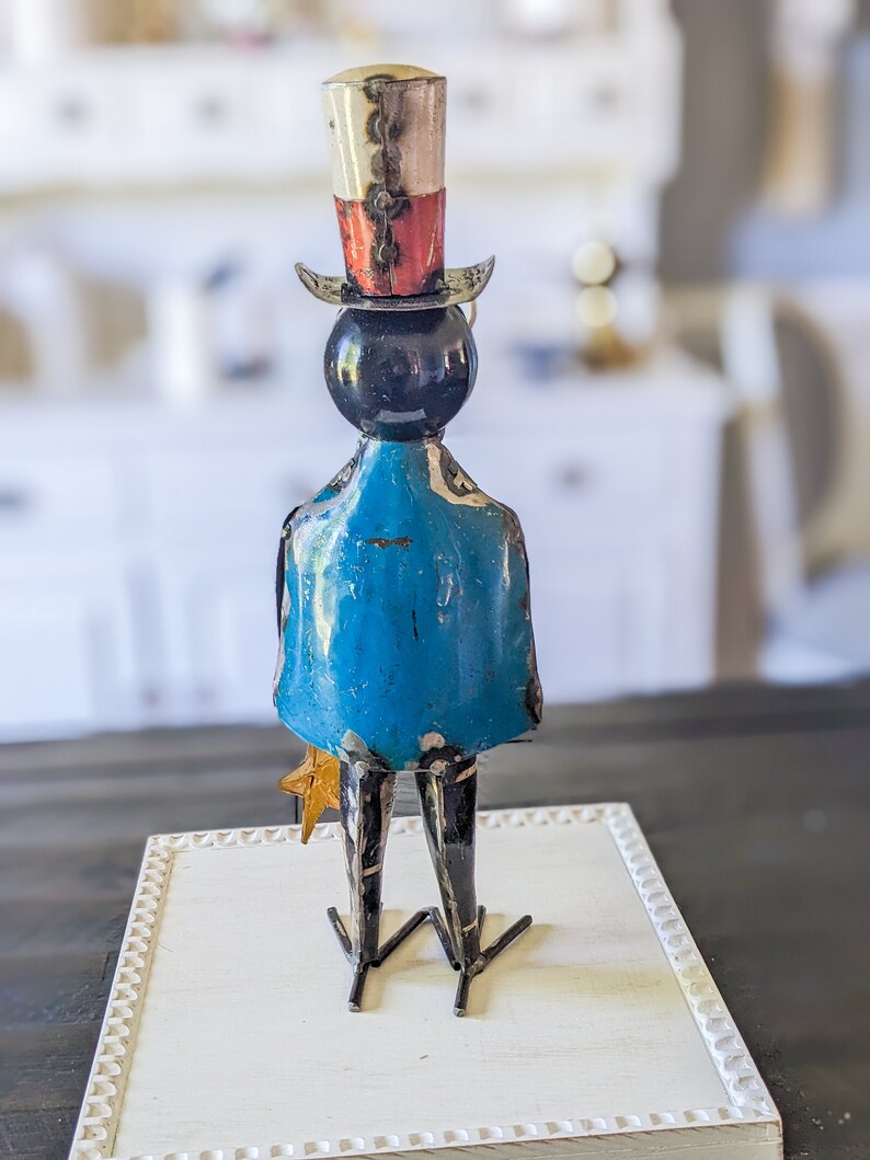 May include: A metal sculpture of a figure with a top hat, blue coat, and star-shaped accessory. The hat is white, red, and silver. The figure is standing on a white, framed surface. The sculpture is a decorative item.