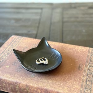 May include: Black ceramic cat-shaped dish with two silver rings inside. The dish is sitting on a brown book with gold lettering and decorative borders.