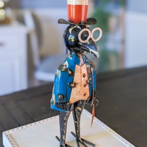 May include: A whimsical metal bird sculpture wearing a top hat, spectacles, and a blue jacket with gold accents. The bird holds a small, red-tipped cane. The sculpture is displayed on a white, decorative tray.
