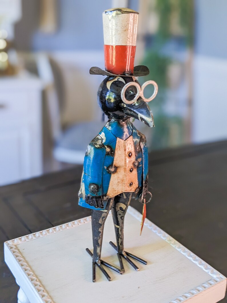 May include: A whimsical metal bird sculpture wearing a top hat, spectacles, and a blue jacket with gold accents. The bird holds a small, red-tipped cane. The sculpture is displayed on a white, decorative tray.