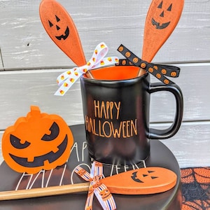 Op de afbeelding: Zwarte koffiemok met de tekst "Happy Halloween" in gouden letters. De mok is omringd door twee oranje houten lepels met pompoengezichten en een kleine oranje pompoen met een zwart gezicht.