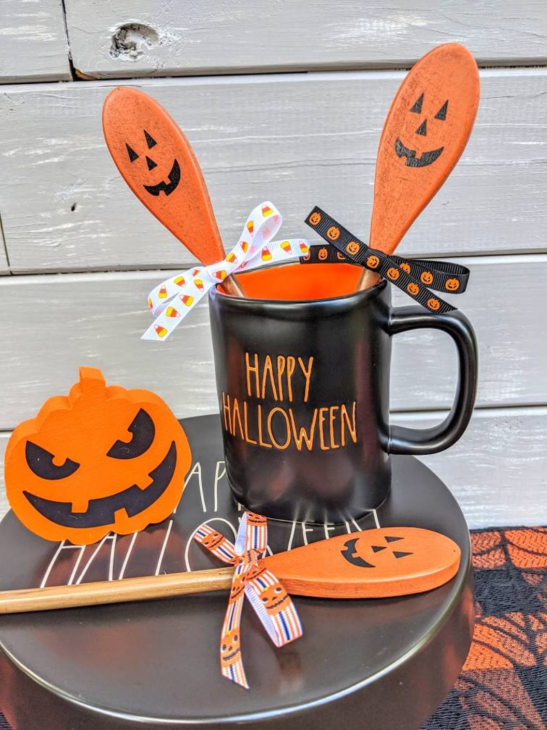 Op de afbeelding: Zwarte koffiemok met de tekst "Happy Halloween" in gouden letters. De mok is omringd door twee oranje houten lepels met pompoengezichten en een kleine oranje pompoen met een zwart gezicht.