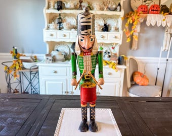 May include: A wooden nutcracker doll with a tall black and silver hat, green jacket, and red drum. The doll has a long white mustache and beard, and black boots. The nutcracker is a classic holiday decoration.