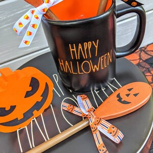 Op de afbeelding: Zwarte keramische mok met oranje tekst die "Happy Halloween" zegt. De mok is versierd met een oranje en wit snoepmais lint en een zwart lint met oranje pompoengezichten. De mok staat op een zwart bord met een pompoenvormige houten lepel en een oranje papieren pompoen-uitsparing.