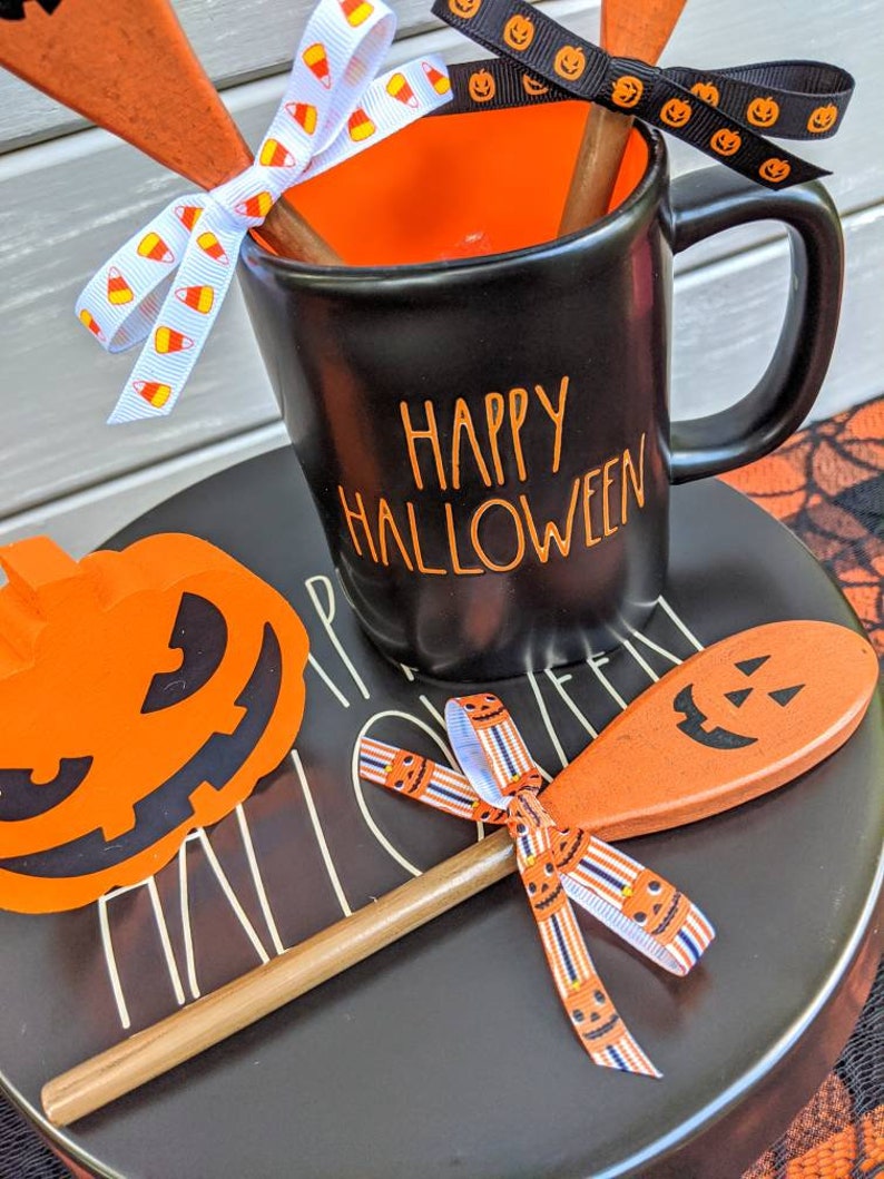 Op de afbeelding: Zwarte keramische mok met oranje tekst die "Happy Halloween" zegt. De mok is versierd met een oranje en wit snoepmais lint en een zwart lint met oranje pompoengezichten. De mok staat op een zwart bord met een pompoenvormige houten lepel en een oranje papieren pompoen-uitsparing.