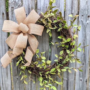 May include: A brown burlap bow tied around a green leafy wreath. The wreath is made of twigs and has a rustic look.