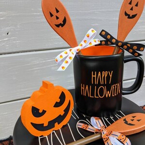 Op de afbeelding: Een zwarte mok met de tekst "Happy Halloween" erop, versierd met oranje en zwart lint en houten lepels met geschilderde pompoengezichten. Een kleine oranje pompoen met een zwart gezicht zit op een zwart bord.
