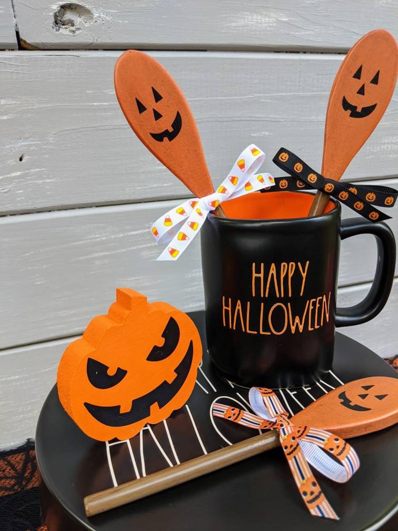 Op de afbeelding: Een zwarte mok met de tekst "Happy Halloween" erop, versierd met oranje en zwart lint en houten lepels met geschilderde pompoengezichten. Een kleine oranje pompoen met een zwart gezicht zit op een zwart bord.