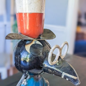 May include: A metal sculpture of a bird wearing a top hat and spectacles. The hat is white and red, and the bird has a black body and blue vest. The sculpture has a vintage, rustic appearance.