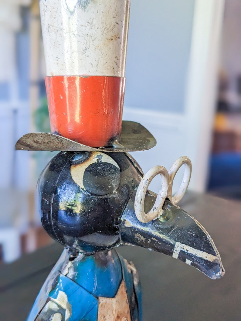 May include: A metal sculpture of a bird wearing a top hat and spectacles. The hat is white and red, and the bird has a black body and blue vest. The sculpture has a vintage, rustic appearance.