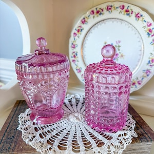 May include: Two pink glass jars with lids, one with a diamond pattern and the other with a hobnail design, sit on a white lace doily atop a vintage book. A floral plate is in the background.