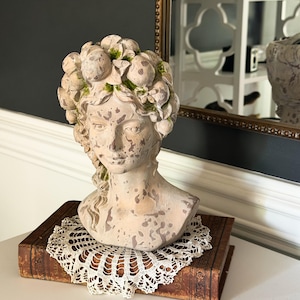 Puede incluir: Un busto de color piedra beige que representa a una persona con un acabado desgastado y envejecido, con toques de musgo verde. La figura tiene una corona de frutas y hojas esculpidas sobre la cabeza, con el cabello trenzado hacia un lado.