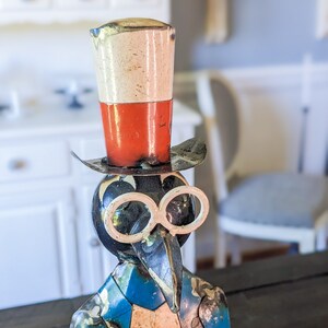 May include: A whimsical metal sculpture of a bird-like figure wearing a top hat and spectacles. The hat is white and red, and the figure has a blue jacket with a white vest. The sculpture has a vintage, handcrafted appearance.