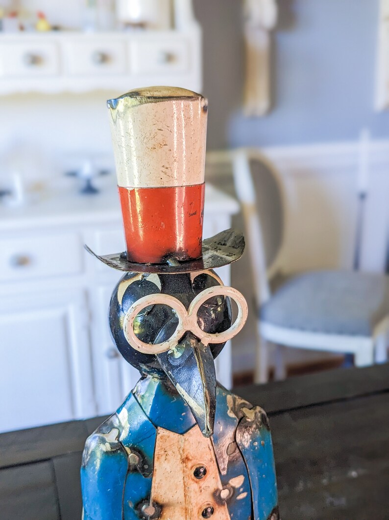 May include: A whimsical metal sculpture of a bird-like figure wearing a top hat and spectacles. The hat is white and red, and the figure has a blue jacket with a white vest. The sculpture has a vintage, handcrafted appearance.