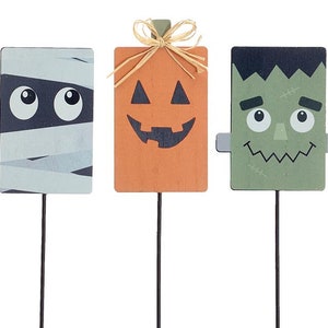 May include: Three Halloween-themed wooden signs with stick stakes. The signs feature a mummy, a pumpkin, and Frankenstein. The mummy is white with black eyes and bandages. The pumpkin is orange with a black jack-o'-lantern face. Frankenstein is green with black hair, stitches, and a wide smile.