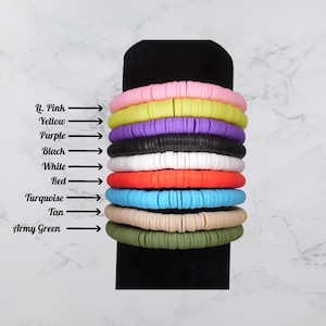 May include: A stack of colorful clay disc bracelets in light pink, yellow, purple, black, white, red, turquoise, tan, and army green. The colors are labeled with arrows. The bracelets are stacked on a black display.