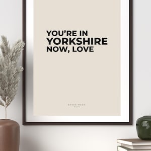 Op de afbeelding: Een ingelijste print met de tekst "YOU'RE IN YORKSHIRE NOW, LOVE" in zwart op een beige achtergrond. De print zit in een donkerbruine lijst en wordt weergegeven op een houten plank. De woorden "BAKER MADE Studio" staan onderaan.