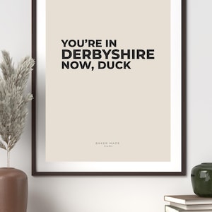Op de afbeelding: Een ingelijste print met de tekst "YOU'RE IN DERBYSHIRE NOW, DUCK" in vette zwarte letters op een beige achtergrond. De print zit in een donkerbruine lijst. De woorden "BAKER MADE Studio" staan onderaan.