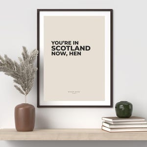 Op de afbeelding: Een ingelijste print met de tekst "YOU'RE IN SCOTLAND NOW, HEN" in zwart op een beige achtergrond. De print zit in een donkerbruine lijst. Een vaas met gedroogde planten en andere decoratieve items staan op een plank eronder.