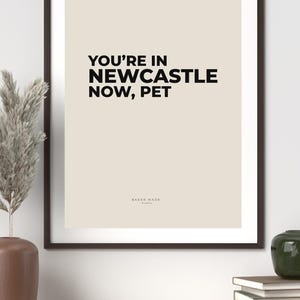 Op de afbeelding: Een ingelijste print met de tekst "YOU'RE IN NEWCASTLE NOW, PET" in zwarte letters op een beige achtergrond. De print zit in een donkerbruine lijst en bevat het logo "BAKER MADE studio".