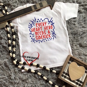 May include: White baby onesie with blue and red text that reads "Every heart hero needs a sidekick". The onesie has a red heart with the text "Little Bow Beauties & More" on it.