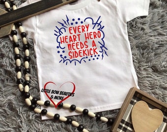 May include: White baby onesie with blue and red text that reads "Every heart hero needs a sidekick". The onesie has a red heart with the text "Little Bow Beauties & More" on it.