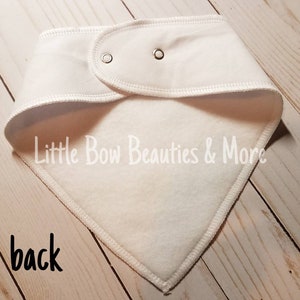 May include: White cotton bandana with a fleece lining. The bandana has two snaps on the back and the text "Little Bow Beauties & More" is printed on the front.