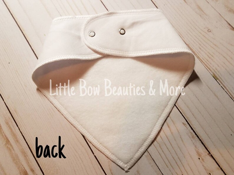 May include: White cotton bandana with a fleece lining. The bandana has two snaps on the back and the text "Little Bow Beauties & More" is printed on the front.