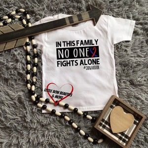 May include: White t-shirt with the text "In this family no one fights alone #CHDWARRIOR" and a blue and red ribbon. The shirt also has the text "Little Bow Beauties & More" and a heart.