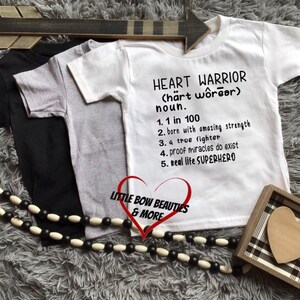 May include: White t-shirt with black text that reads "HEART WARRIOR (härt wôrear) noun. 1. 1 in 100 2. born with amazing strength 3. a true fighter 4. proof miracles do exist 5. Real life SUPERHERO".