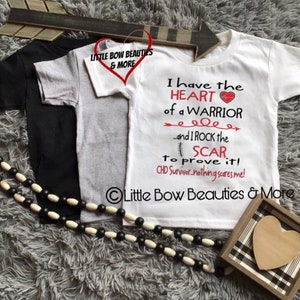 May include: Two white t-shirts with a red heart and text that reads "I have the HEART of a WARRIOR...and I ROCK the SCAR to prove it! CHD Survivor-nothing scares me!"  The text is on a white t-shirt with a black and white checkered heart in a frame below the text.  The t-shirts are on a gray fuzzy surface with a black and white beaded necklace.