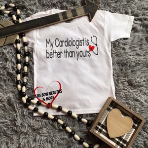 May include: White t-shirt with the text "My Cardiologist is better than yours" and a red heart with a stethoscope. The shirt has a small logo that says "Little Bow Beauties & More".