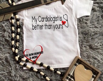 May include: White t-shirt with the text "My Cardiologist is better than yours" and a red heart with a stethoscope. The shirt has a small logo that says "Little Bow Beauties & More".