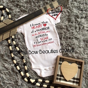 May include: White baby onesie with the text "I have the HEART of a WARRIOR...and I ROCK the SCAR to prove it! CHD Survivor-nothing scares me!"  A red heart is next to the word "WARRIOR". The onesie is on a gray fuzzy surface with a black and white beaded necklace and a wooden box with a black and white plaid design and a wooden heart inside.