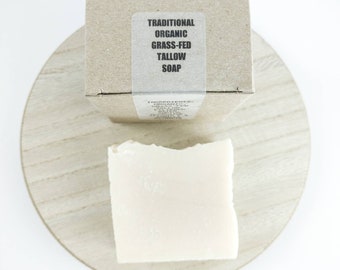May include: A bar of white soap on a wooden surface. The soap is packaged in a brown cardboard box with a label that reads "Traditional Organic Grass-Fed Tallow Soap".