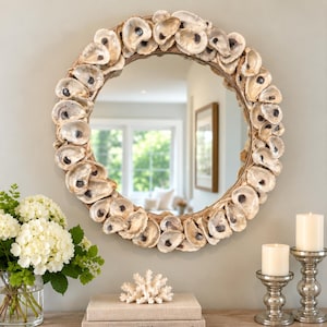 May include: A circular wall mirror framed with a dense arrangement of natural oyster shells. The shells feature varied shades of cream, beige, and grey, creating a textured, layered border around the reflective glass surface of the round mirror.
